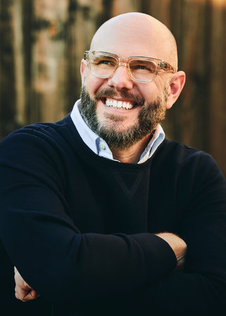 A picture of author Steven Rowley in a dark navy sweater with a light blue button up shirt underneath. His arms are folded across his chest and he's smiling and looking up to the left. He has on clear glasses and a beard. He is bald. He is on a brown blurred background.