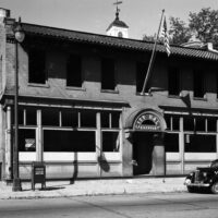 A black and white photograph shows the City Hall of Kirkwood with an American flag flying above the entrance. A vintage car is parked on the street in front of the building.