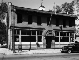 A black and white photograph shows the City Hall of Kirkwood with an American flag flying above the entrance. A vintage car is parked on the street in front of the building.