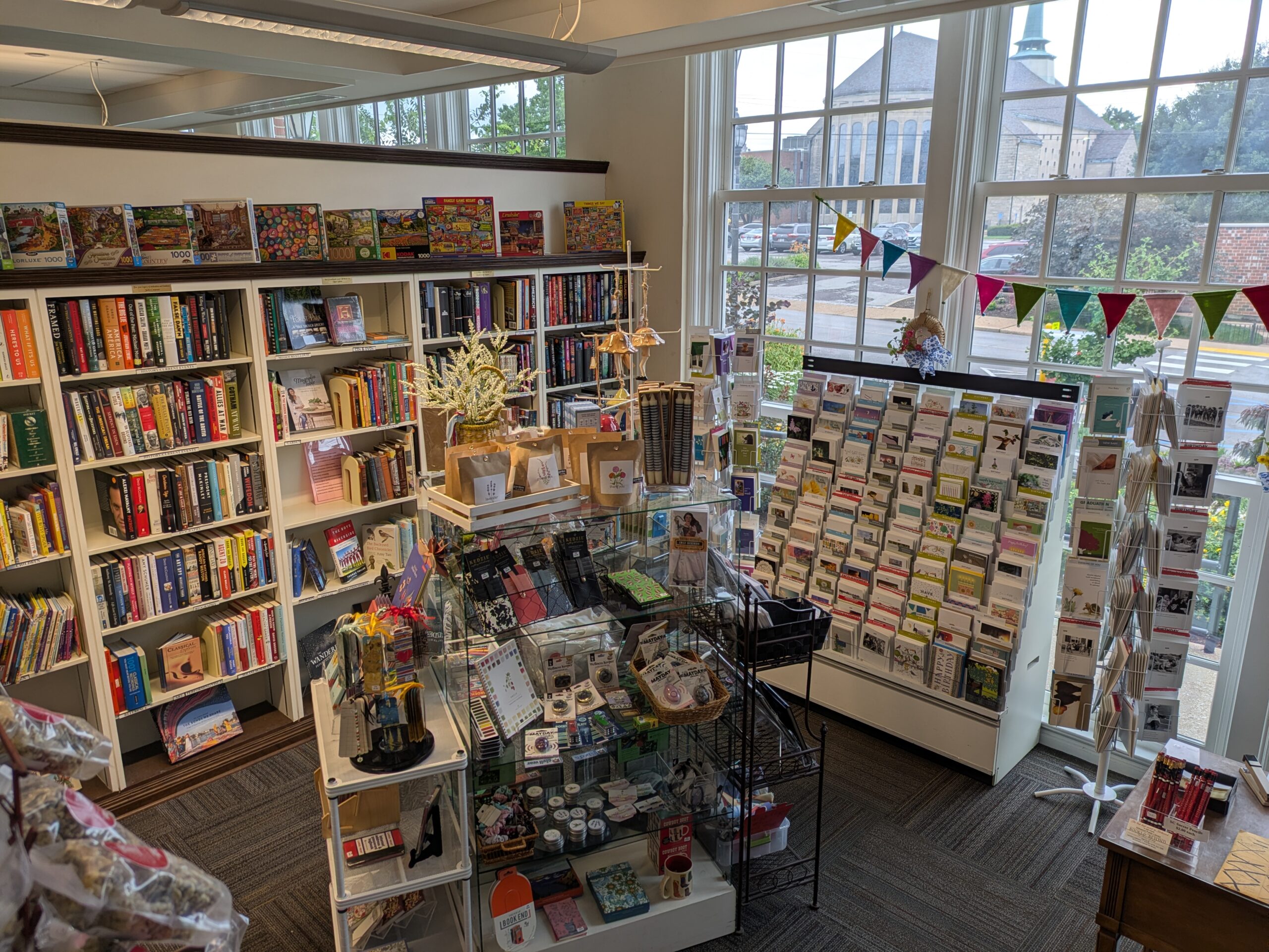 Books-and-Beyond A view of a bookstore interior featuring shelves packed with books, a display of greeting cards, and a large window overlooking a street with buildings.